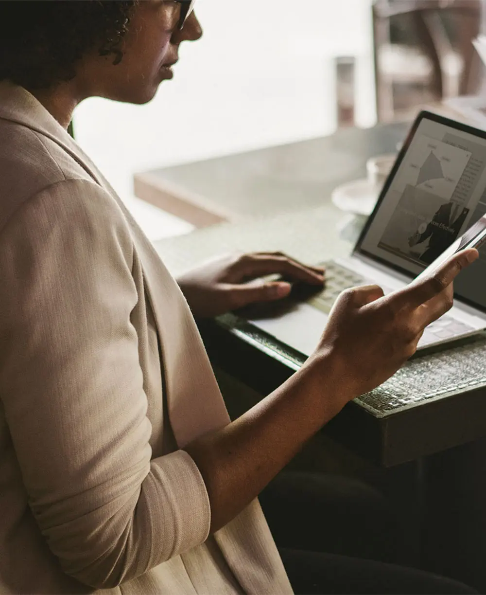 An image of a woman working on a laptop and holding a phone in her hand.