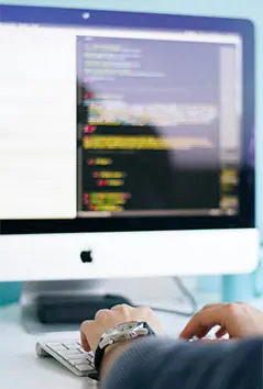 An image of a person's hands typing on a keyboard with a blurred apple computer in the background that has a blurred out screen filled with programming code.