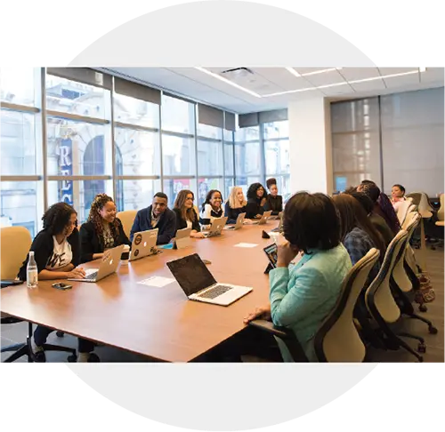 An image of a boardroom with people working at a large table, laptops and paper covering the table.