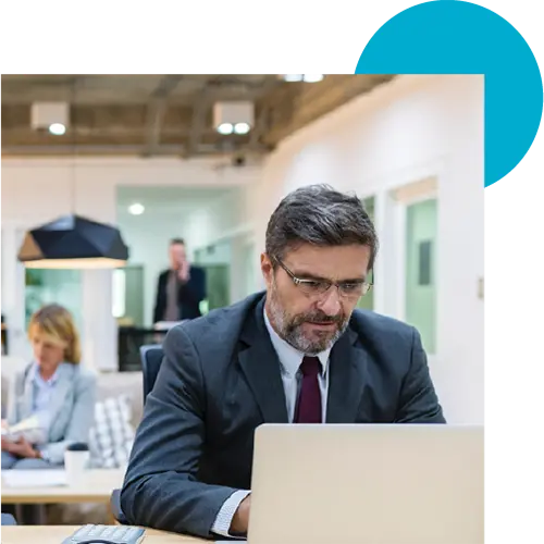 An image of a middle aged man with glasses in a suit working on a laptop in an office.